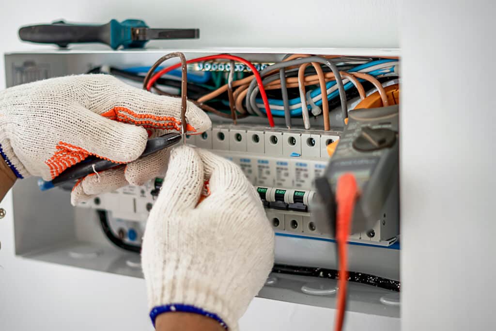 Installation tableau électrique : électricien câble disjoncteurs Électricien en gants blancs manipulant des fils colorés dans un tableau électrique. Des disjoncteurs affichent "O-OFF". Un outil et multimètre visibles.