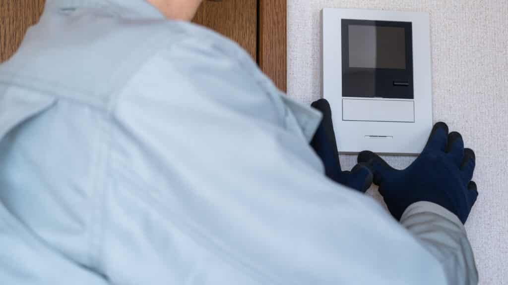 Installation interphone vidéo : technicien expert en action Technicien ganté en uniforme installant un interphone vidéo blanc sur un mur texturé près d'un cadre de porte en bois.