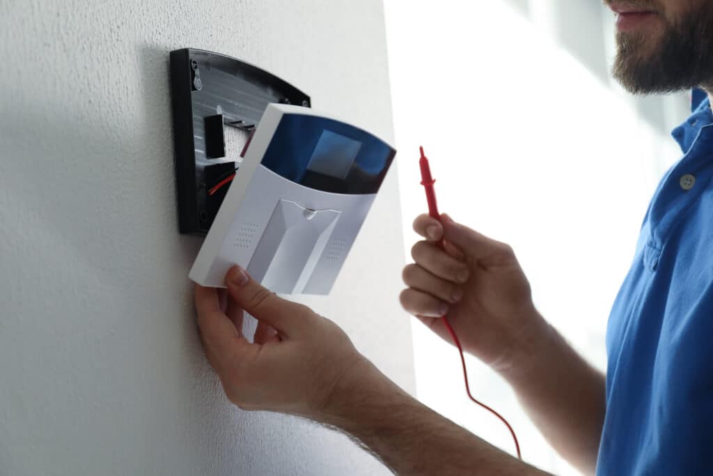 Installation système d'alarme : sécurité maison facile Homme barbu en polo bleu installant un système d'alarme blanc sur un mur texturé, tenant une sonde rouge pour vérification.