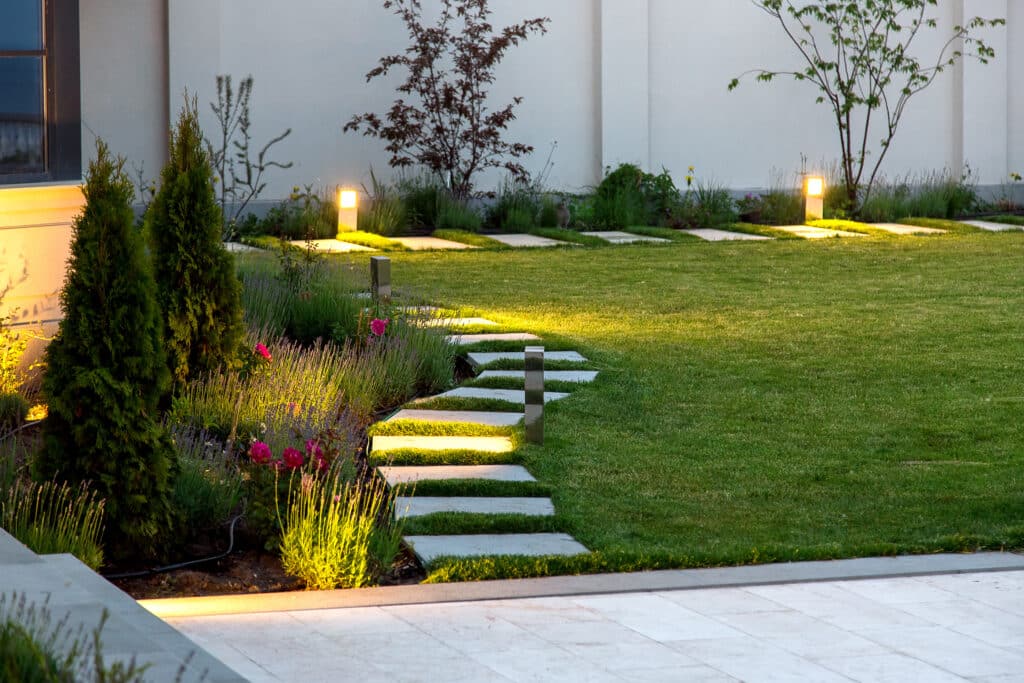 Éclairage de jardin moderne : chemin lumineux et verdure la nuit Jardin moderne avec chemin éclairé la nuit. Lampes intégrées aux dalles, lavande, roses, et thuyas. Un chat observe près du mur.