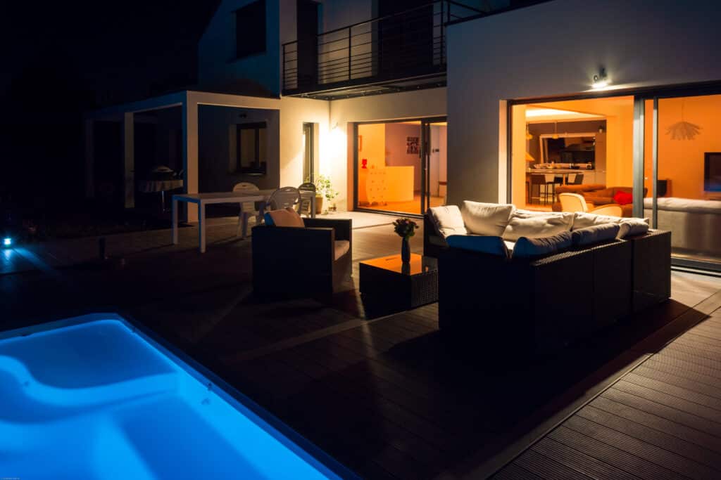 Maison moderne avec piscine et terrasse | Nuit élégante Villa contemporaine la nuit, patio en bois, piscine bleue lumineuse, salon de jardin et intérieur chaudement éclairé.